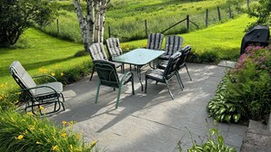 Une terrasse extérieure avec une table et des chaises, entourée d’une végétation luxuriante et d’une pelouse bien entretenue.