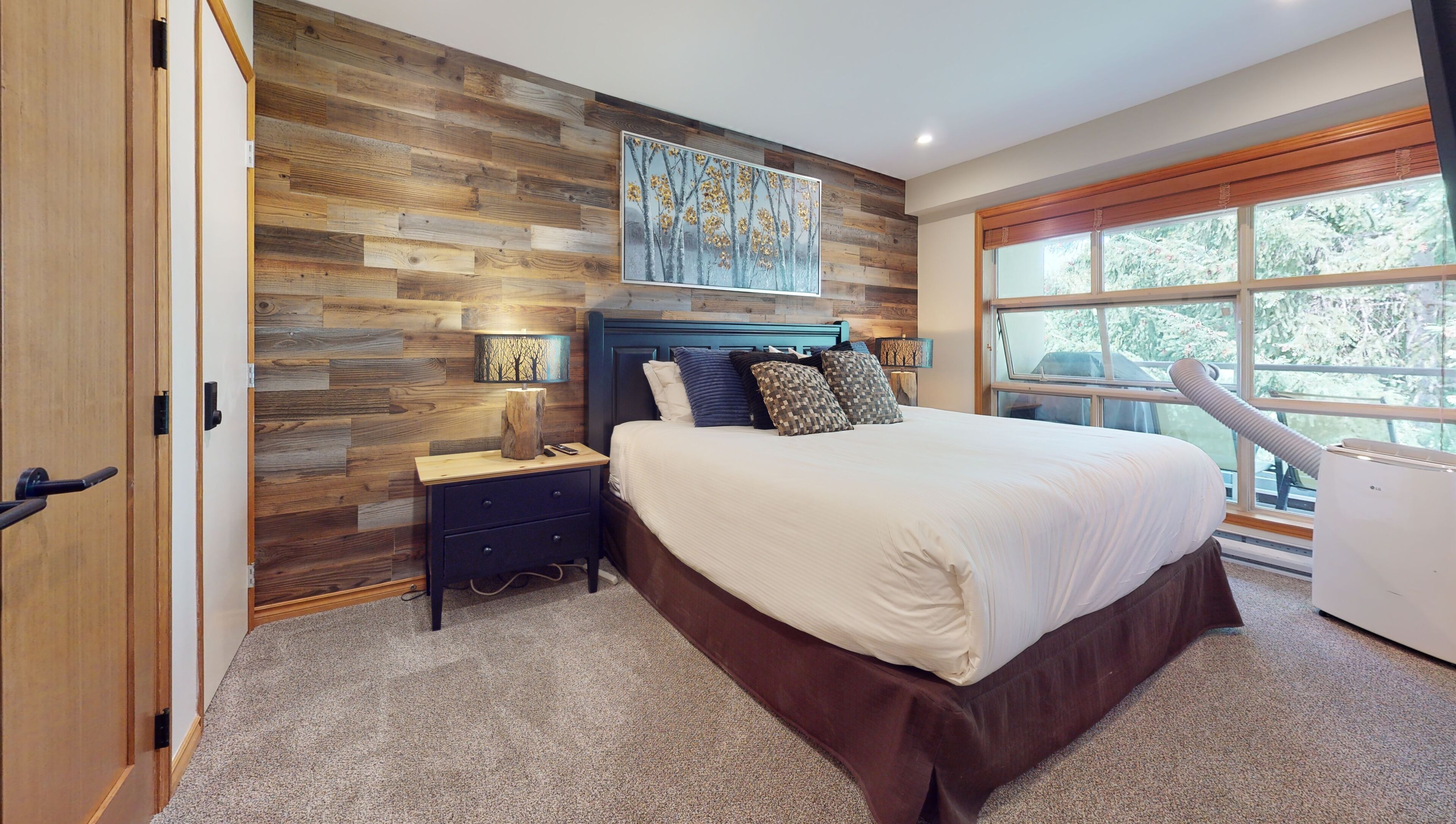 A bedroom with a wooden wall panel, a bed with white bedding, a nightstand, a lamp, and a window with a view of trees.
