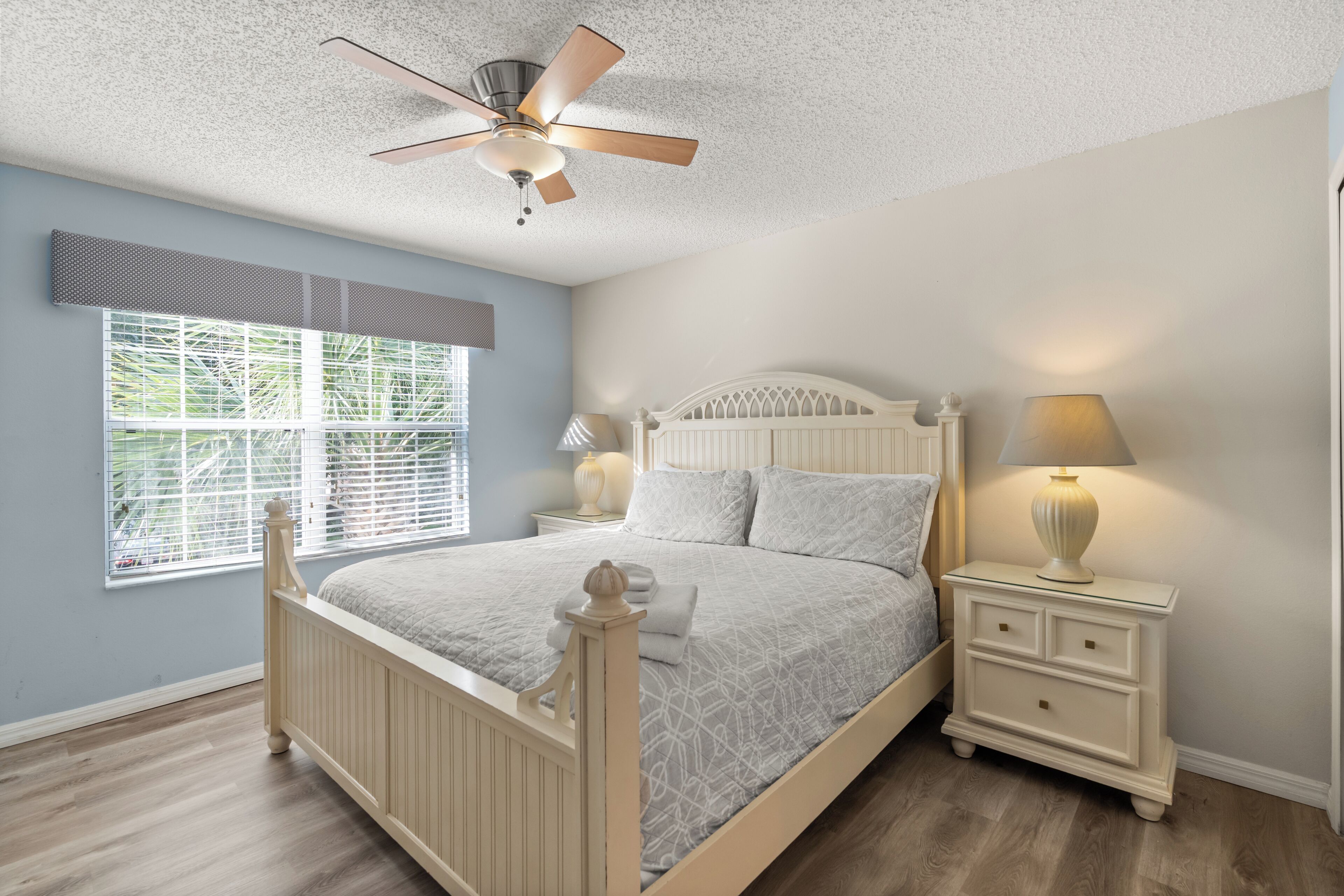 Une chambre à coucher avec un lit, des tables de chevet, un ventilateur de plafond et une fenêtre avec des stores.