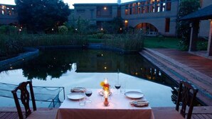 Una mesa de comedor preparada para dos con un centro de mesa de vela, con vista a un estanque tranquilo y un jardín ajardinado.