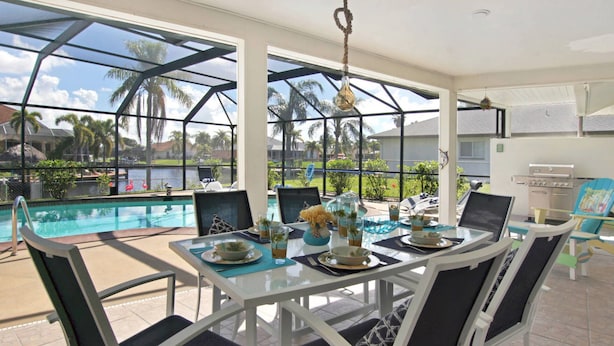 Un espace repas aménagé au bord de la piscine, avec une table et des chaises en verre, offrant une vue sur la piscine et les palmiers.