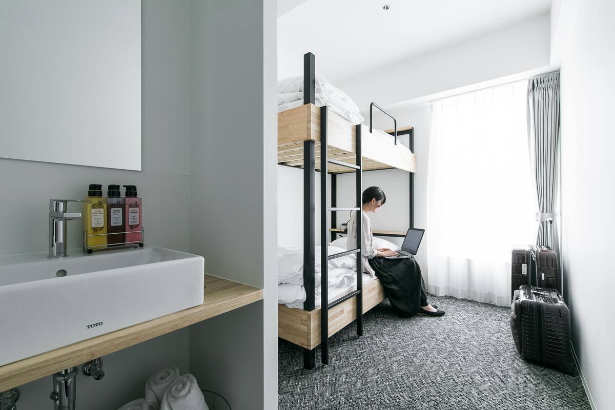 A hotel room with a bunk bed, a sink with toiletries, a laptop, and luggage.