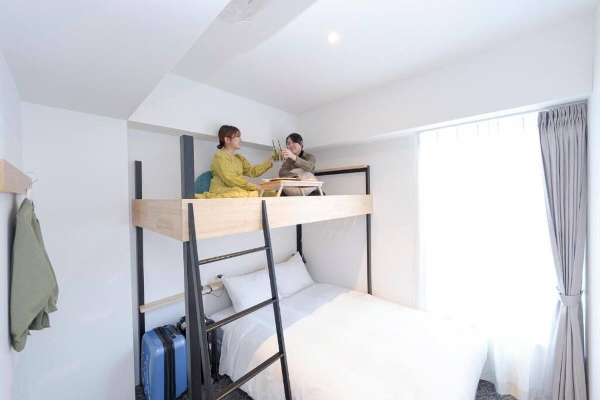 A bunk bed room with a desk and chairs, a suitcase, and a window with curtains.
