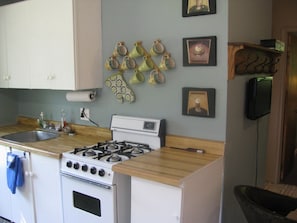 Une cuisine avec des meubles blancs, une cuisinière à gaz et un évier. On y trouve également des éléments décoratifs sur les murs, notamment des tableaux encadrés et un téléviseur fixé au mur.