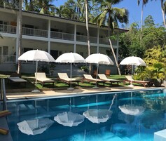 Zona de piscina con sillones de descanso y sombrillas, un edificio con balcones y palmeras al fondo.