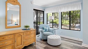 Une pièce comprenant une commode en bois, un fauteuil bleu, un pouf rond, un grand miroir et une fenêtre donnant sur un paysage verdoyant.