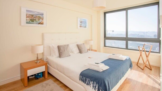 A modern hotel room with a large bed, a nightstand, a window with a city view, and artwork on the walls.