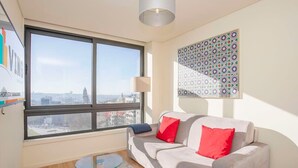 A modern living room with a large window, a grey sofa with red cushions, a glass coffee table, and a patterned wall decoration.