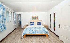 A bedroom with a bed, two bedside tables, a window with curtains, and a hallway leading to another room.