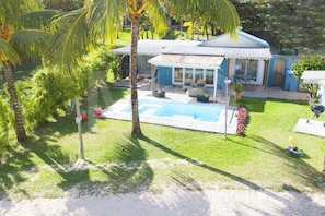 Una casa con piscina, un patio con mobiliario exterior y un jardín con palmeras.
