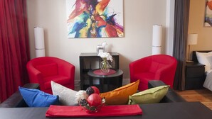 Een moderne kamer met twee rode fauteuils, een zwarte salontafel met decoratieve objecten, een kleurrijk abstract schilderij aan de muur en een bed met wit beddengoed.