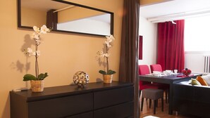 Een hotelkamer met een donker houten dressoir, twee potorkideeën, een decoratieve bol en een zithoek met rode stoelen en een tafel gedekt voor twee personen.