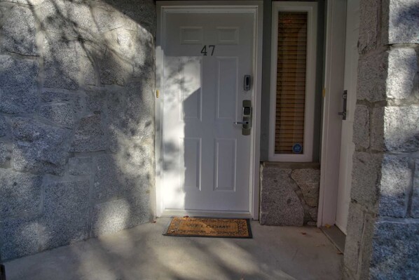 A white door with the number 47, a stone wall, a small window with blinds, and a doormat.