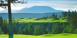 Een golfbaan met glooiende heuvels, bomen en bergen op de achtergrond.