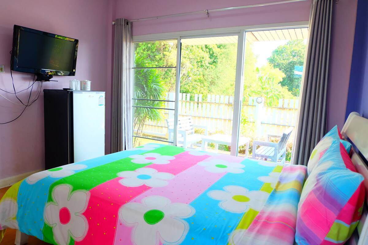 A bedroom with a colorful bedspread, a television mounted on the wall, a refrigerator, and a view of a fenced outdoor area through large windows.