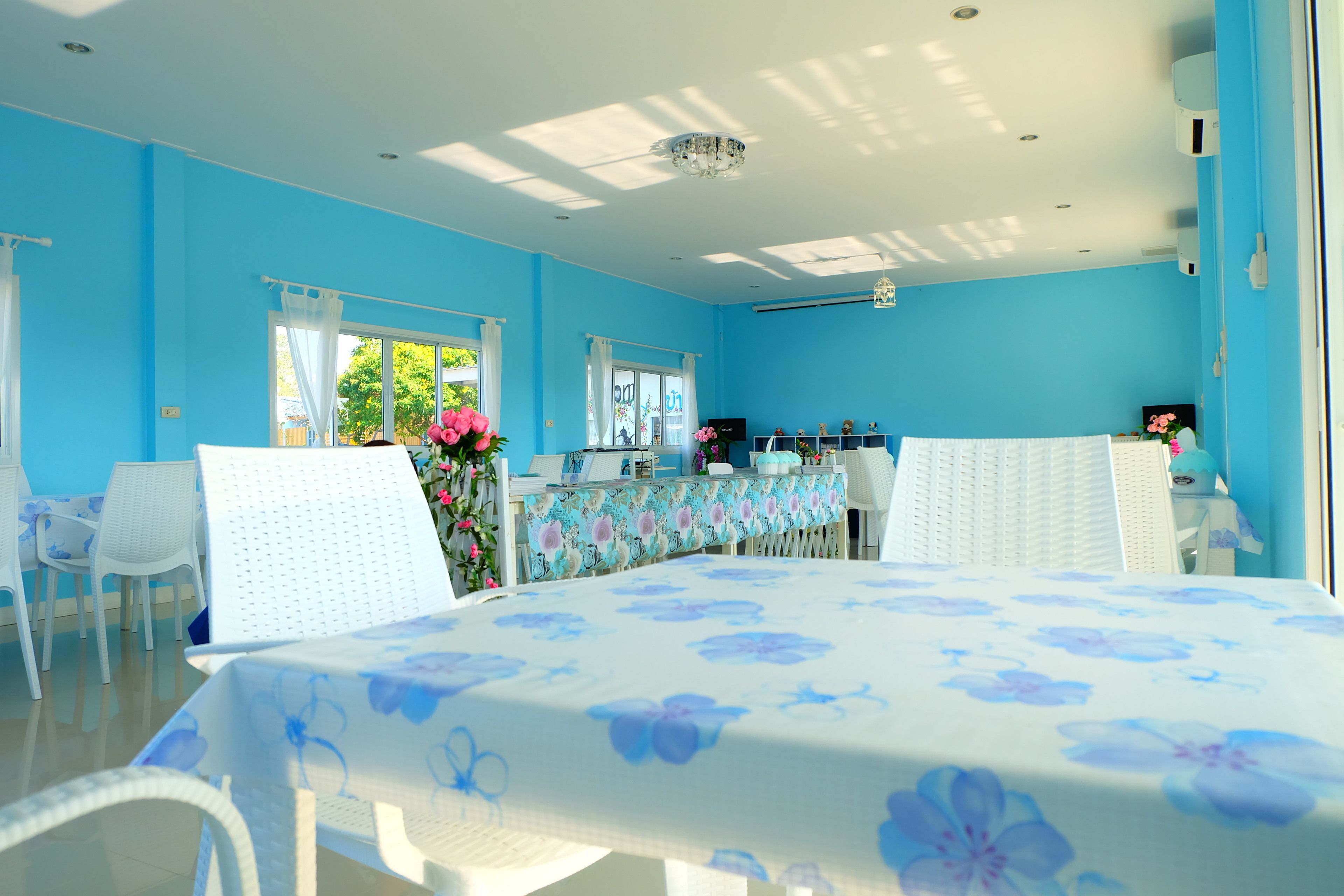 Um quarto claro com paredes azuis, uma mesa com toalha de mesa florida, cadeiras brancas e uma janela com vista para o verde.