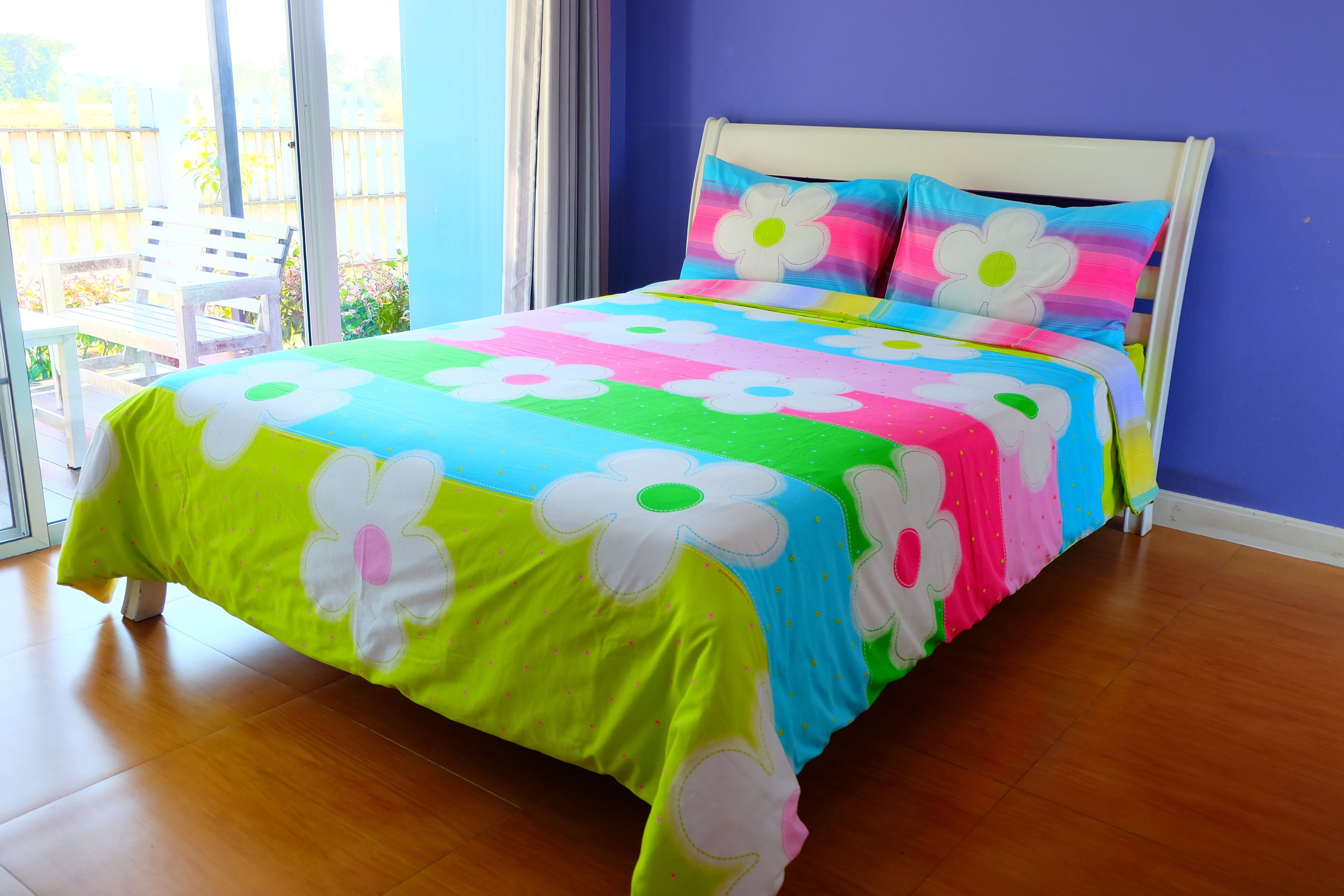 Un letto con una coperta e cuscini dai colori vivaci e motivi floreali, una testiera bianca e vista su un balcone con una sedia bianca.