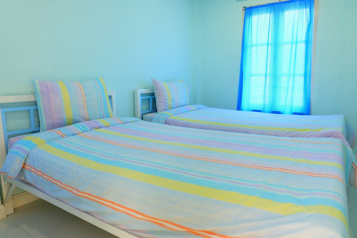 A room with two beds, each with striped bedding, a window with blue curtains, and a light blue wall.