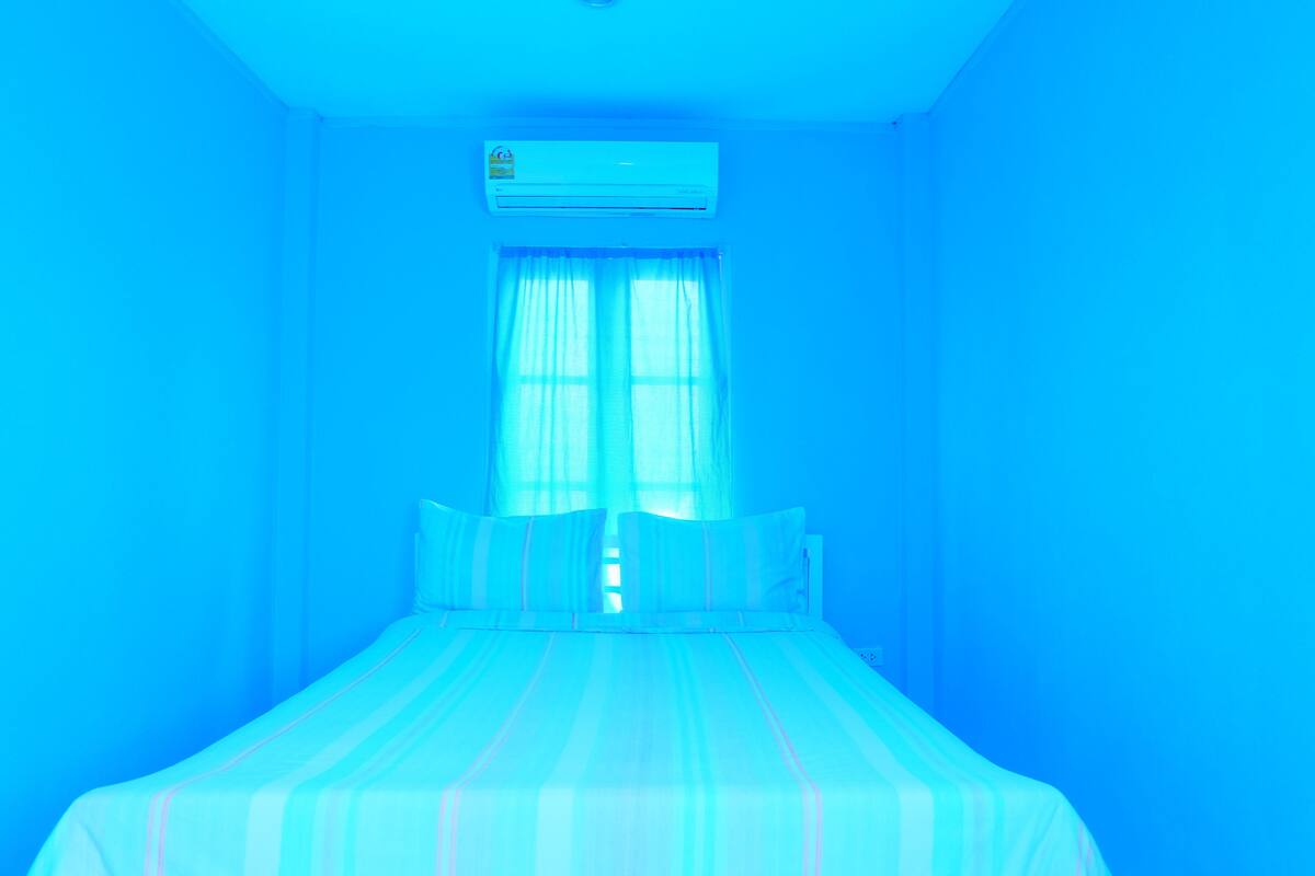 A room with a bed, a window with sheer curtains, and an air conditioning unit on the wall.