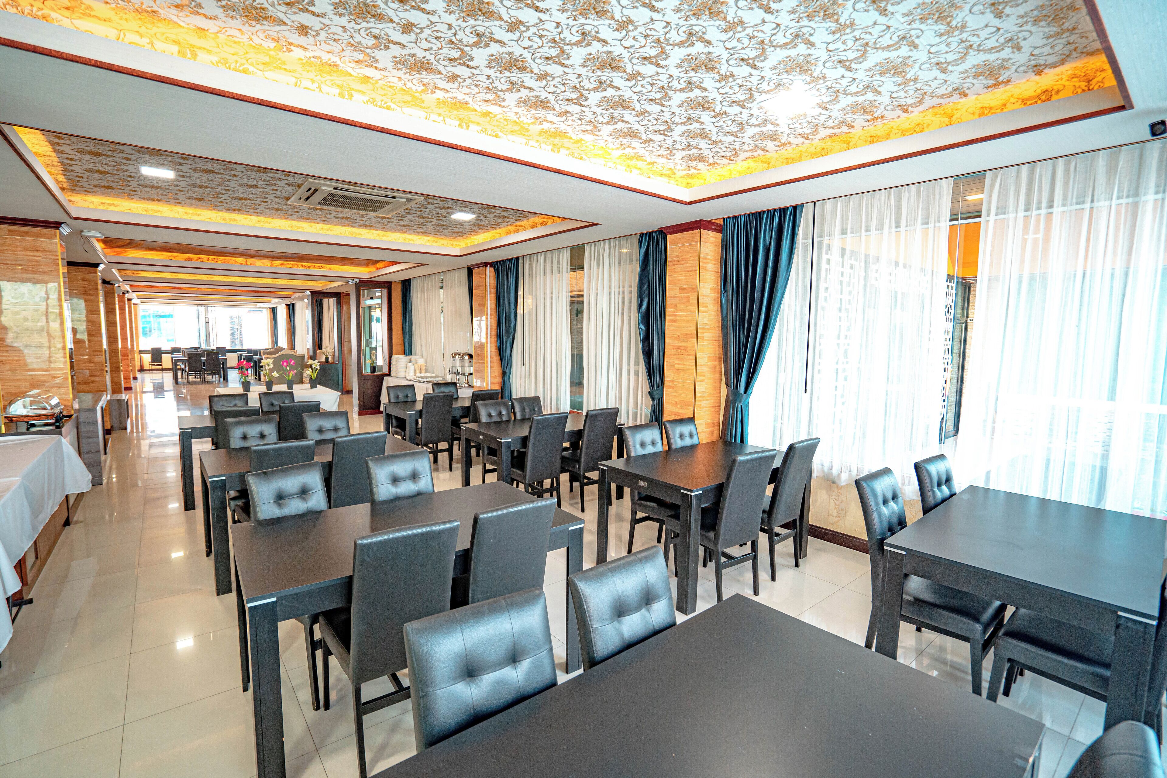 A hotel dining area with multiple tables and chairs, a patterned ceiling, and large windows with curtains.
