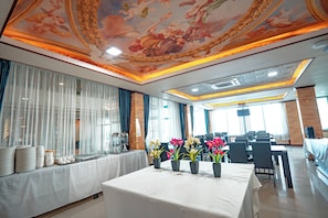 Une salle à manger d’hôtel avec une longue table dressée pour un repas, un plafond orné de motifs décoratifs et une vue sur la ville à travers de grandes fenêtres.