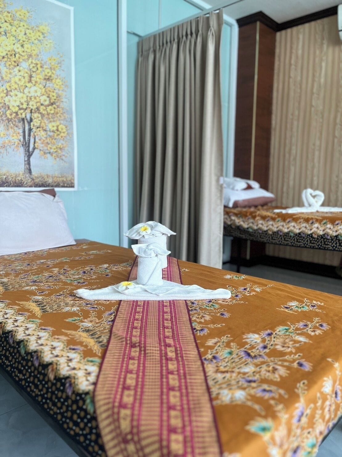 Una habitación de hotel con una cama cubierta por una colcha de motivos florales, un mantel individual blanco con una flor y un cuadro de un árbol en la pared.