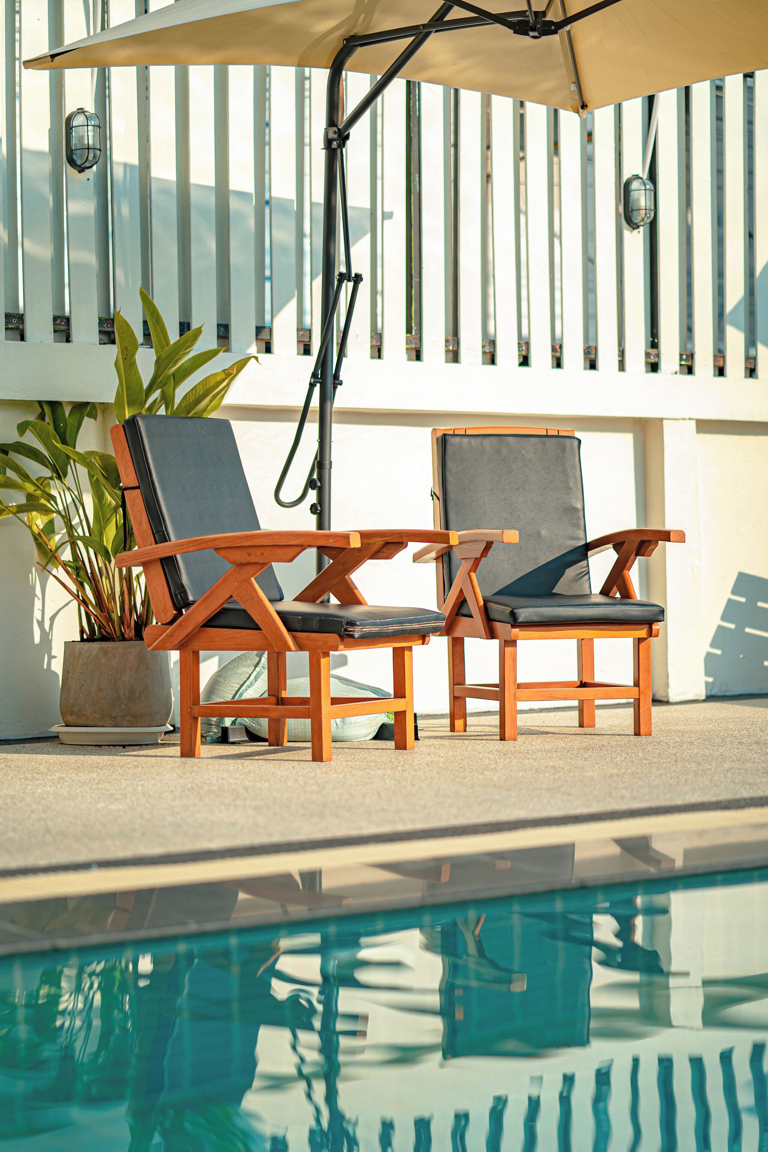 A poolside area with two wooden lounge chairs, a potted plant, and a pool.