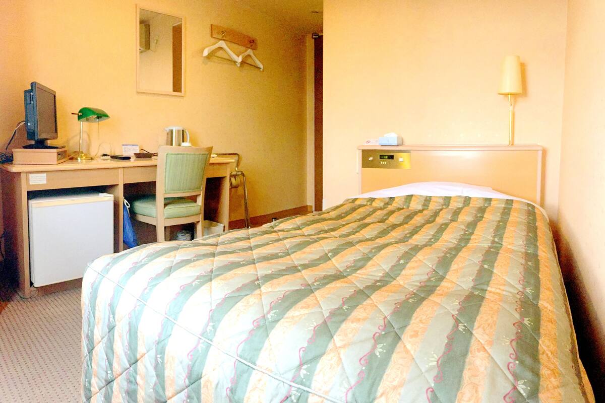 Habitación de hotel con cama, escritorio con computadora, silla, refrigerador pequeño, espejo y mesita de noche con lámpara.