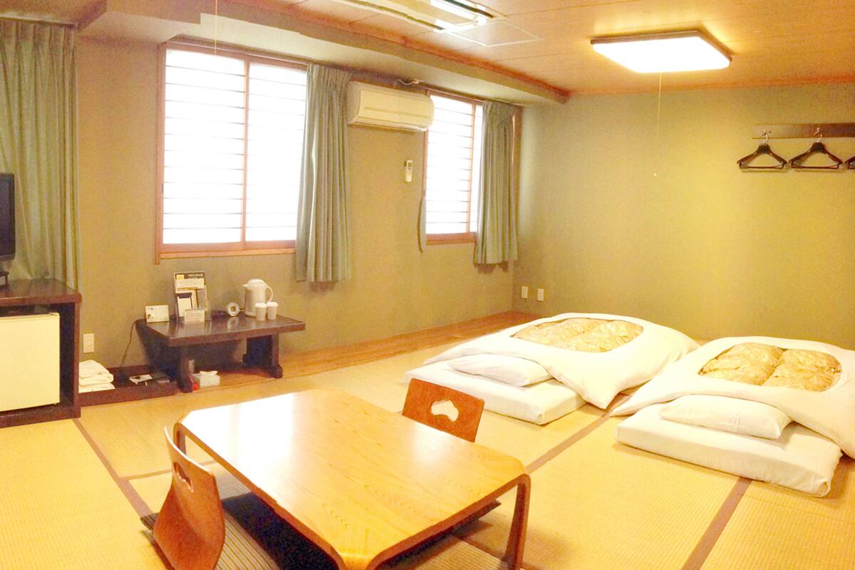 Habitación de hotel con una mesa de madera, dos camas con ropa blanca, un escritorio con teléfono y una televisión.