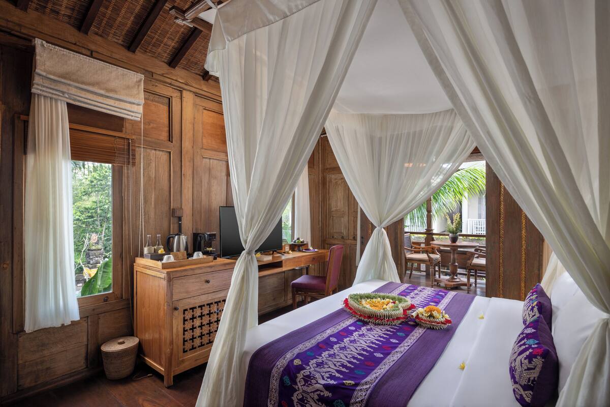 A room with a bed, a desk, and a chair. There is a window with curtains and a view of greenery outside.