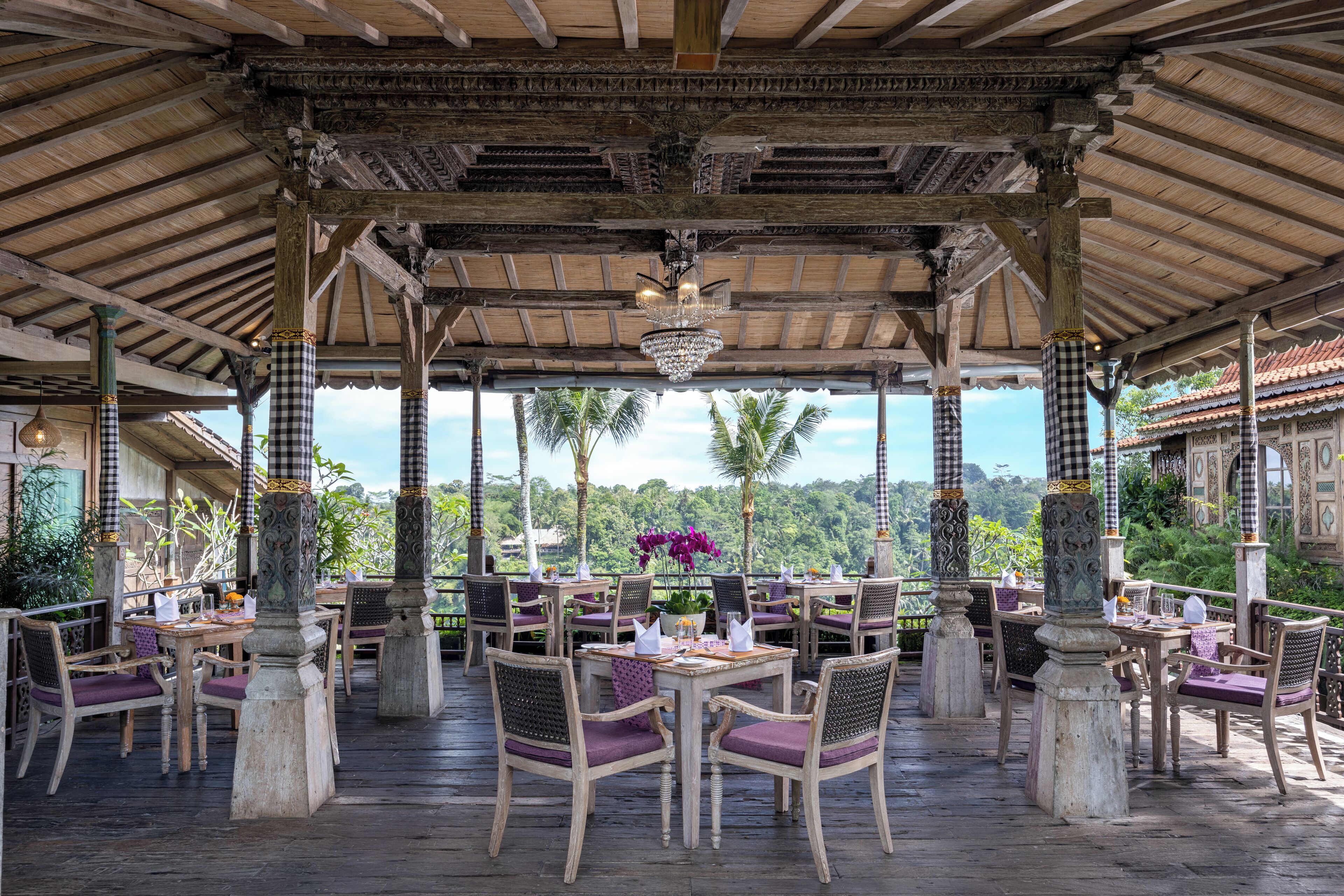 An outdoor dining area with checkered pillars, wooden tables, and purple chairs, overlooking a lush landscape.