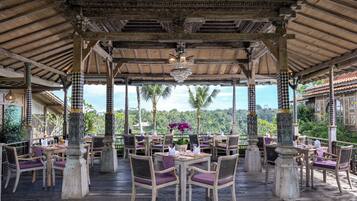 An outdoor dining area with checkered pillars, wooden tables, and purple chairs, overlooking a lush landscape.