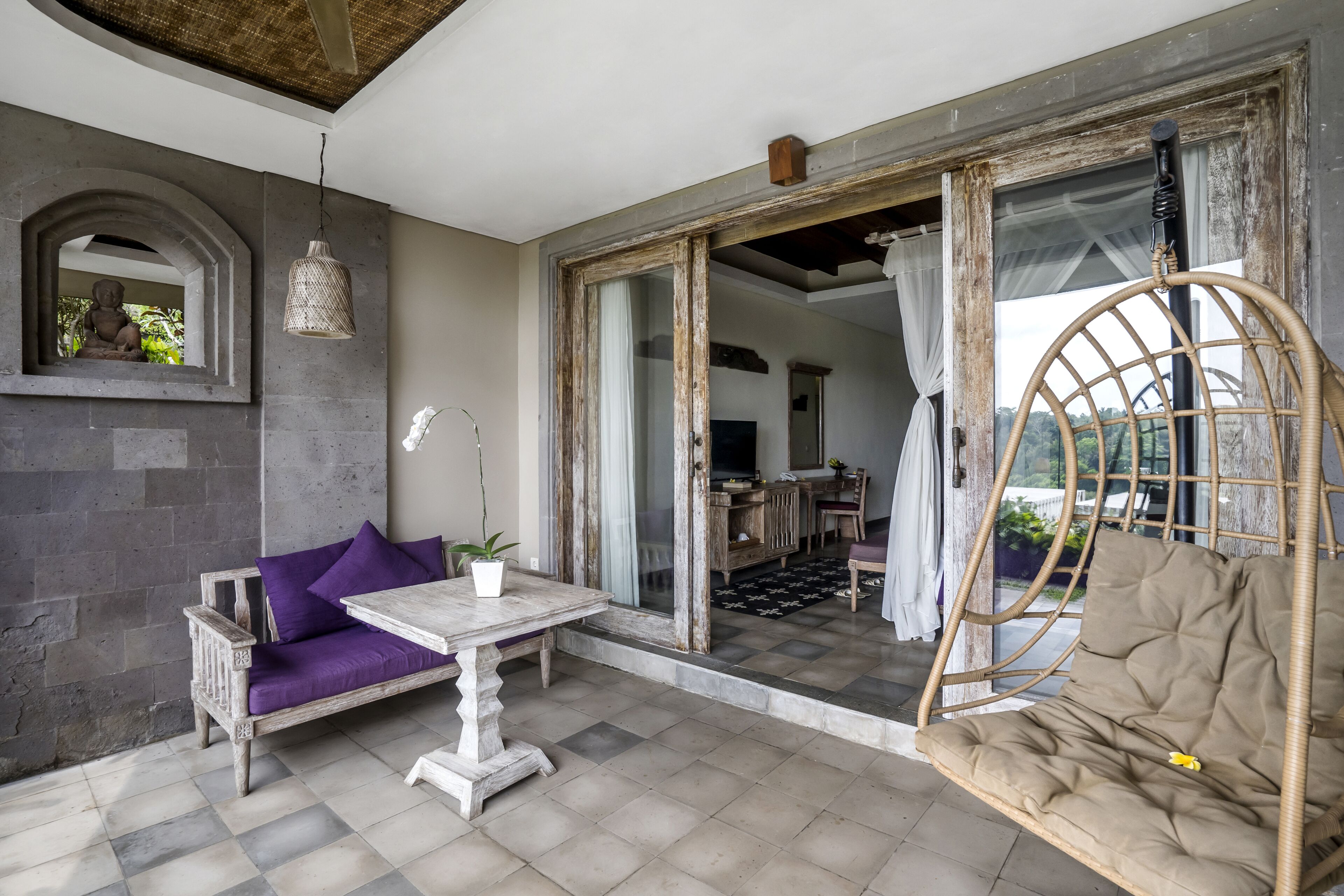A room with a sofa, table, and wicker swing chair. There is a view of a dining area through a glass door.