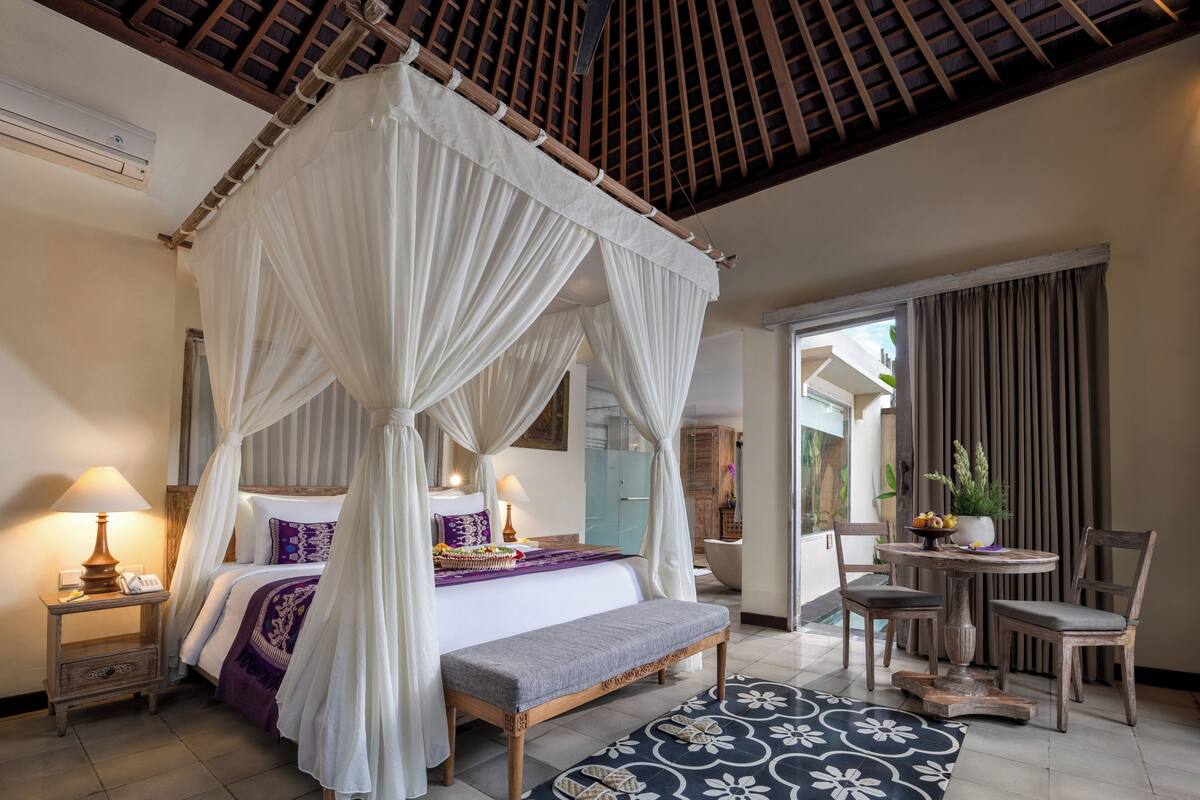 A bedroom with a four-poster bed, bedside tables, a bench, a dining area with chairs, and a view of the outside.