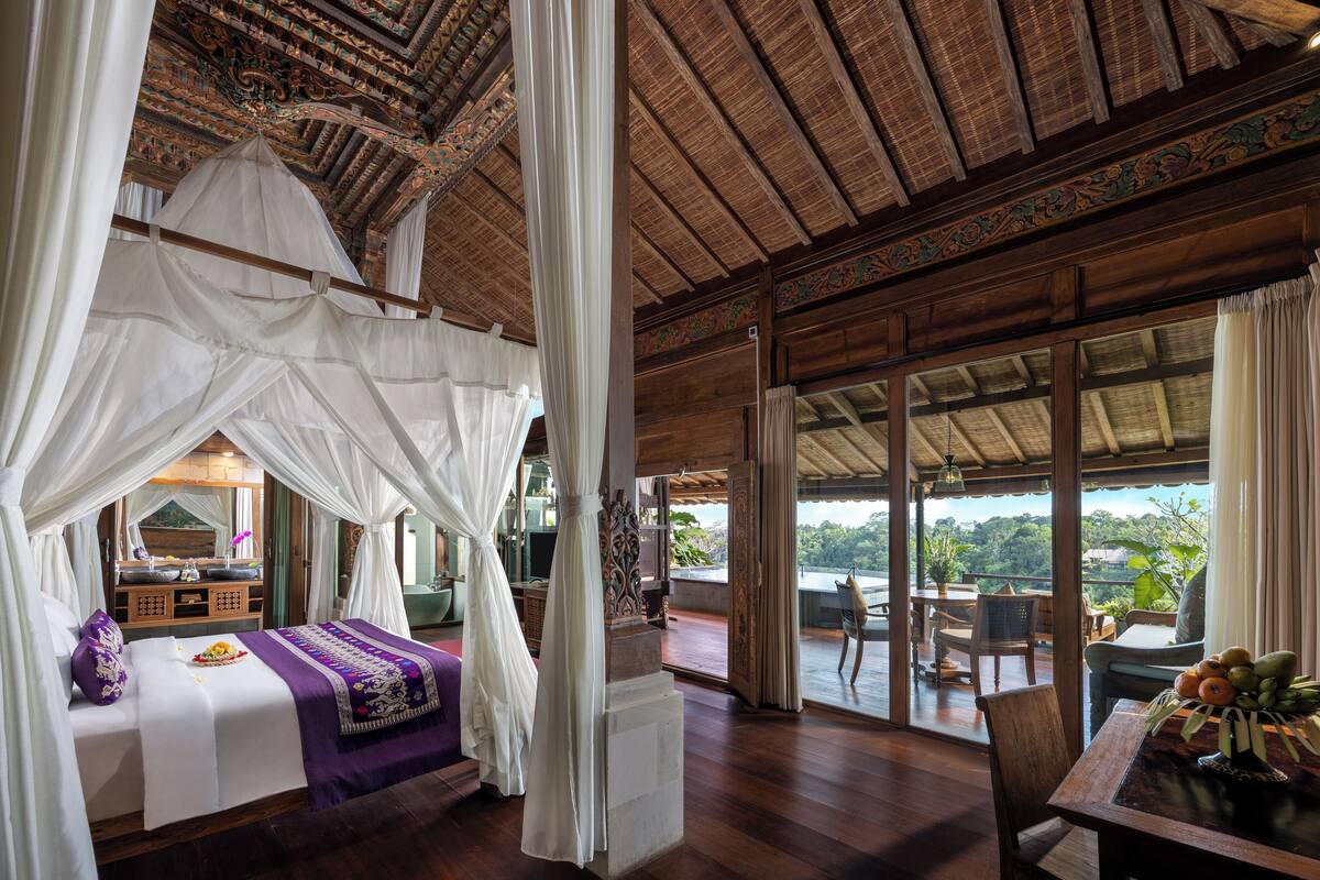 A room with a bed under a canopy, a balcony with a view, and a dining area with a table and chairs.