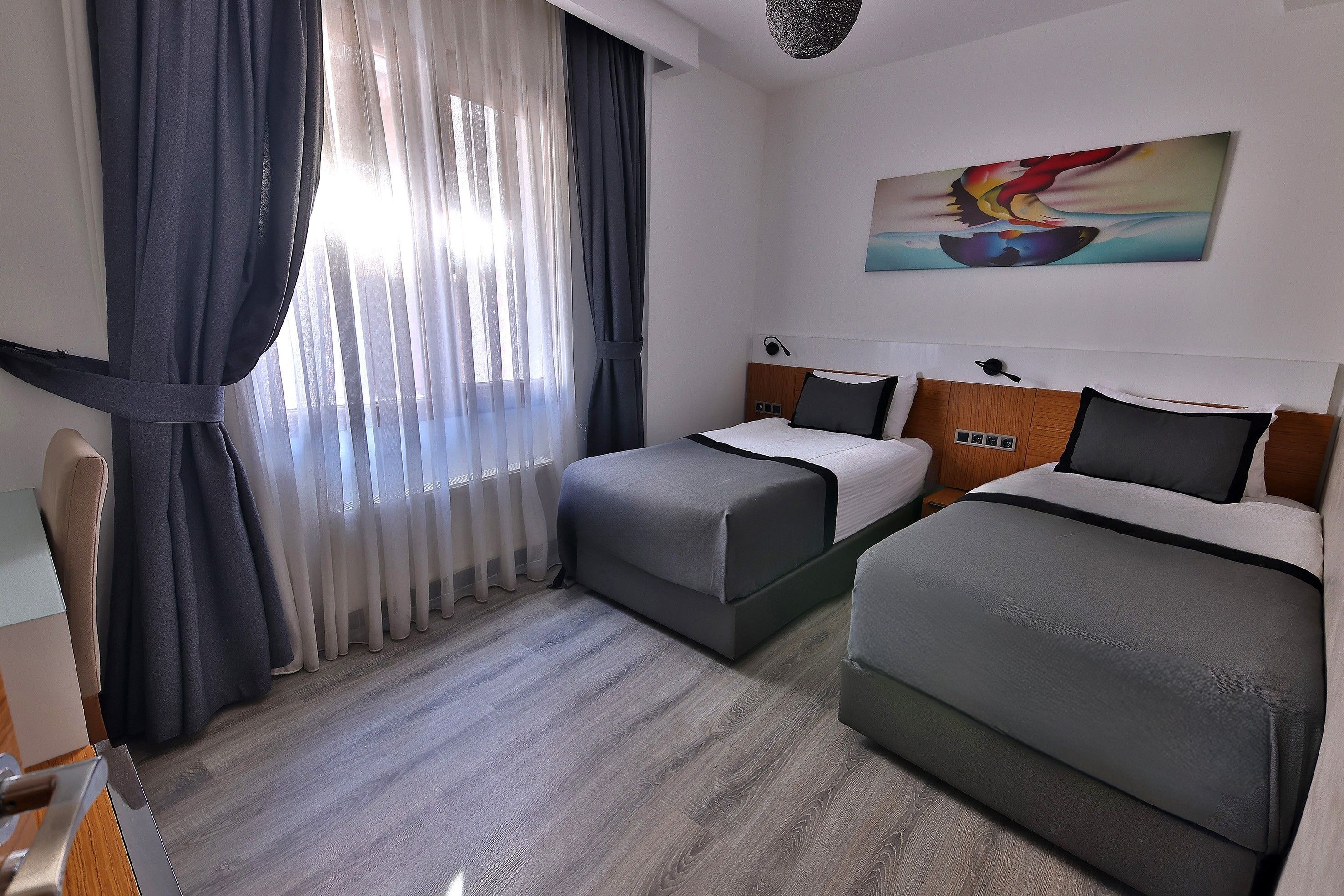 Habitación de hotel con dos camas, un ventanal con cortinas, un cuadro en la pared y suelo de madera.