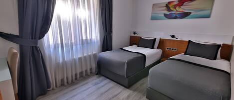 A hotel room with two beds, a large window with curtains, a painting on the wall, and a wooden floor.