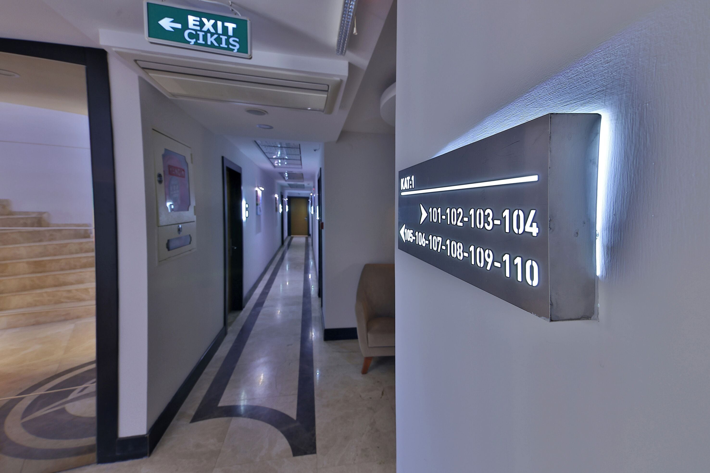 A hotel hallway with an exit sign, a directory sign listing room numbers, and a staircase.