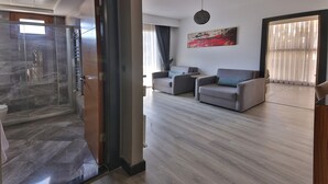A modern living room with a glass shower enclosure, a grey sofa set, and a large abstract painting on the wall. - BUCA MAT HOTEL BY TAHTALI (Izmir)