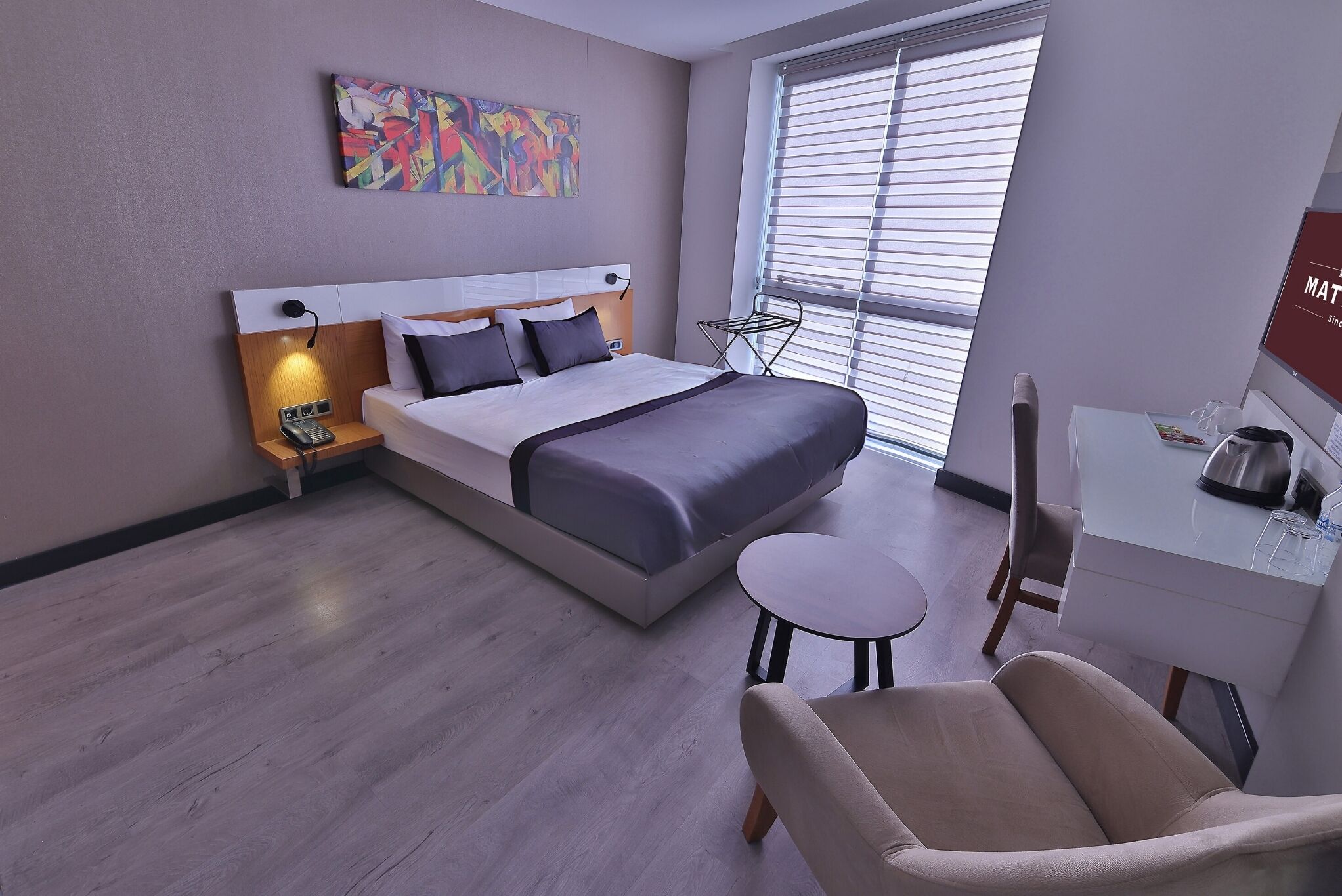 A modern hotel room with a bed, bedside table, lamp, chair, desk, and window with blinds.