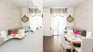 A dining area with a wooden table set for two, white chairs, and a sofa. There are two wall clocks and a window with sheer curtains.