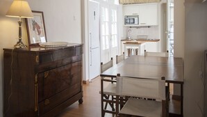 A dining area with a wooden table and chairs, a dark wooden dresser with a lamp, and a kitchen in the background.