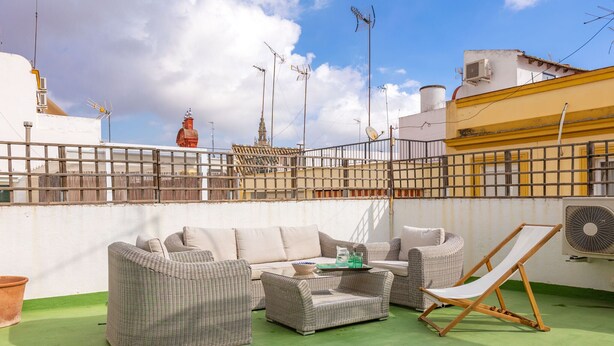 A rooftop terrace with wicker furniture, a fan, and a view of the city skyline.
