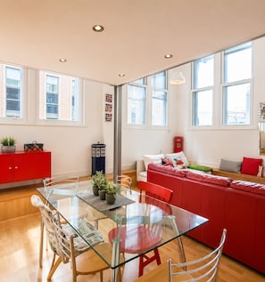 A modern living space with a glass dining table, red furniture, and large windows providing natural light. - 1 LIVERPOOL LOFT (liverpool)