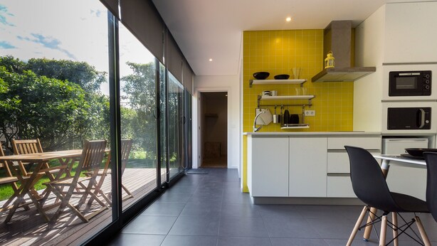 Een moderne keuken met een groot raam, een eethoek met een houten tafel en stoelen, en een gele spatwand.