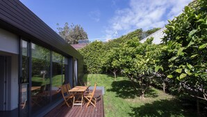 Een modern huis met een houten terras, een glazen deur naar een patio, en een weelderige tuin met diverse planten en bomen.