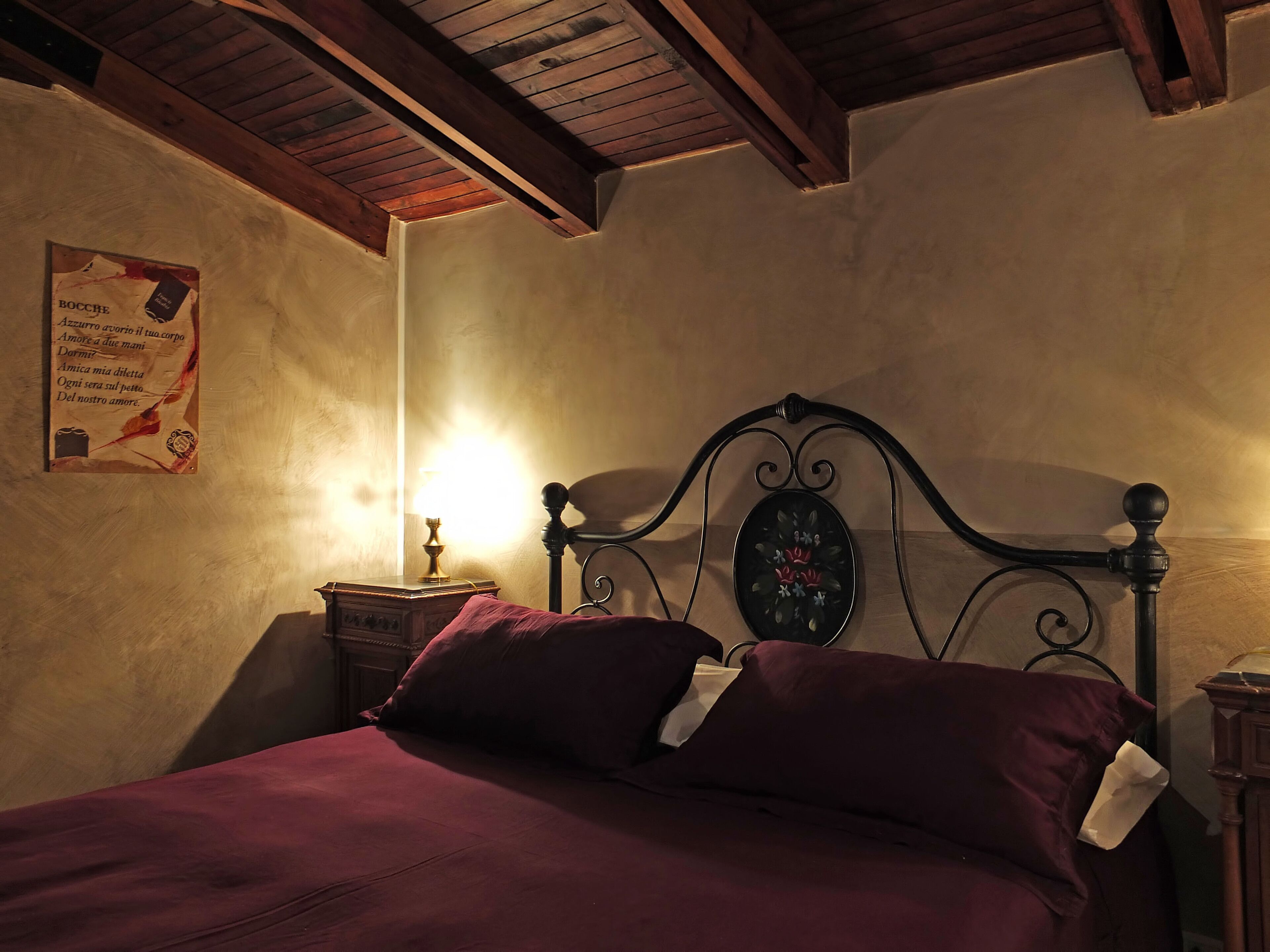 Une chambre avec un plafond en bois, un lit doté d’une tête de lit ornée de motifs floraux, une table de chevet avec une lampe et une affiche accrochée au mur.