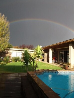 A backyard with a swimming pool, a house, and a rainbow in the sky.