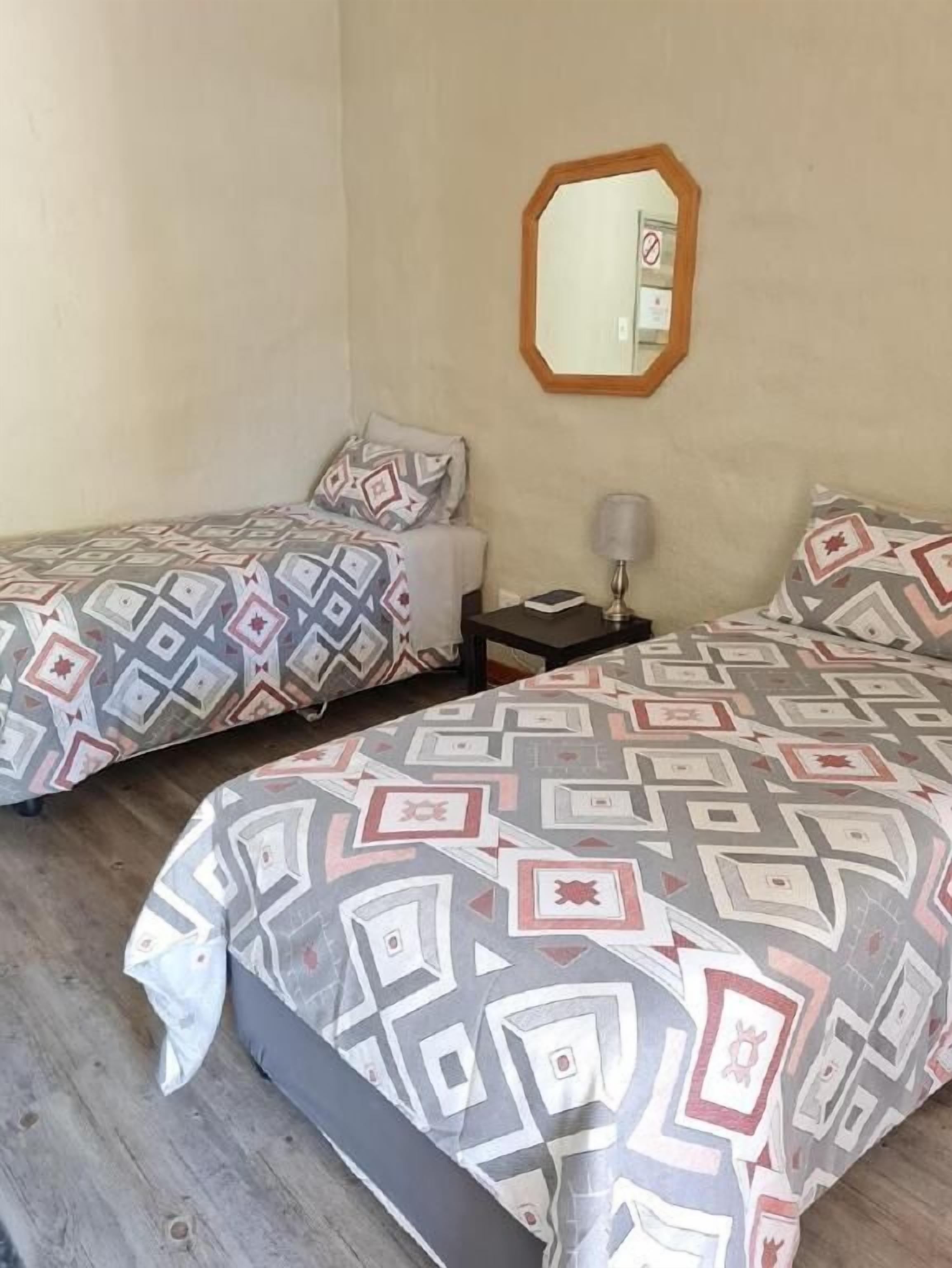 A room with two beds, each with patterned bedding, a nightstand with a lamp, and a mirror on the wall.