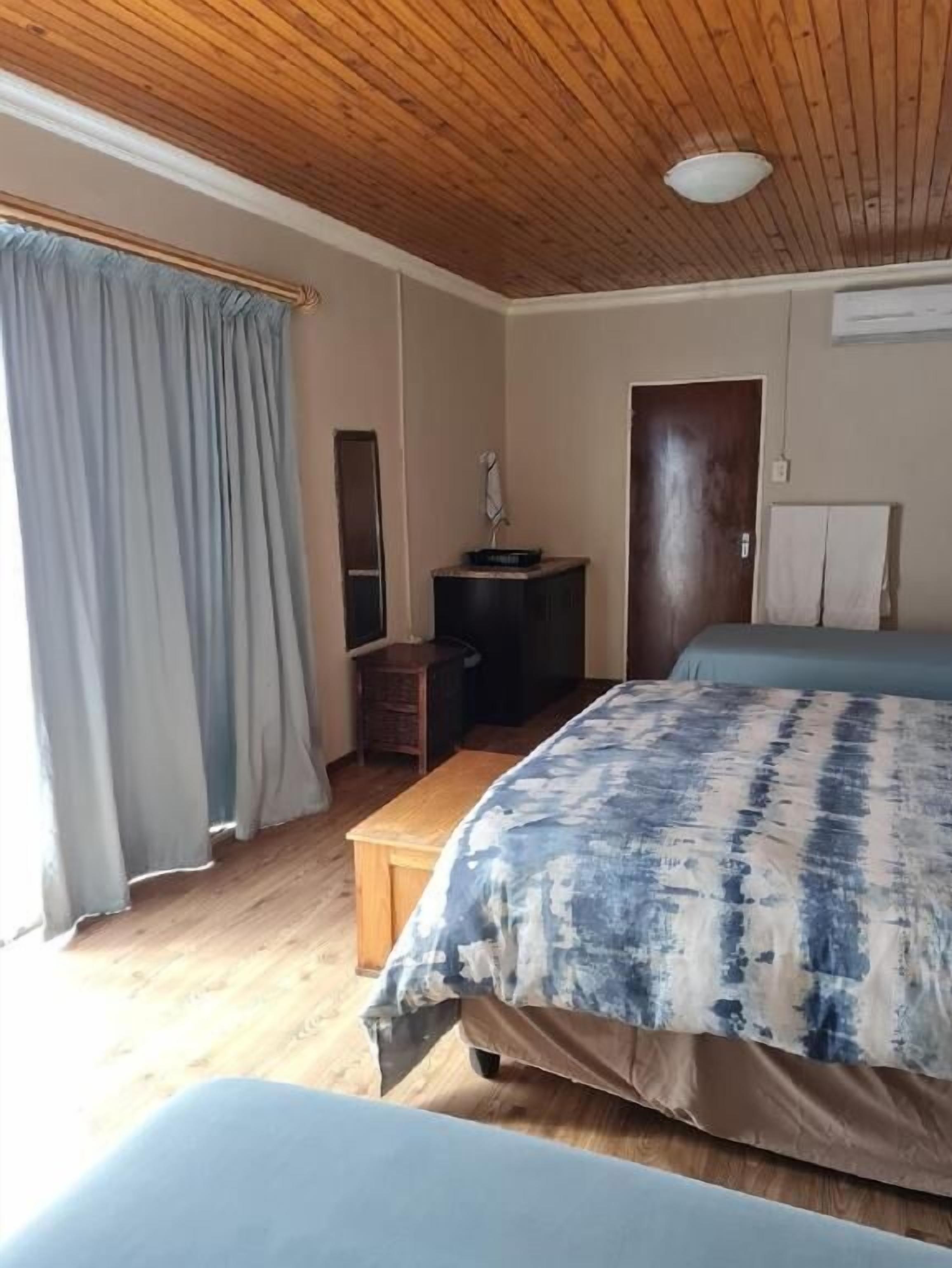 Un dormitorio con techo de madera, una cama con colcha azul y blanca, un banco de madera, una pequeña mesa con una lámpara, y una puerta con una toalla colgada.